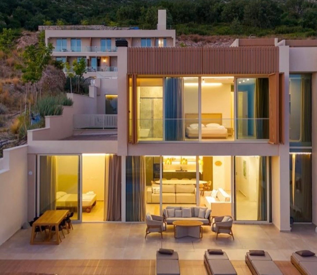 Modern hillside home with illuminated interior, large glass doors opening to patio with lounge chairs at dusk.