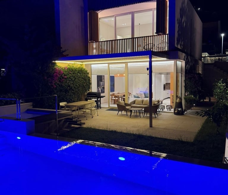 Modern two-story house with illuminated interior and patio overlooking a glowing blue swimming pool at night