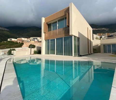 Modern villa with turquoise swimming pool, wooden and cream facades, mountainous backdrop under cloudy sky