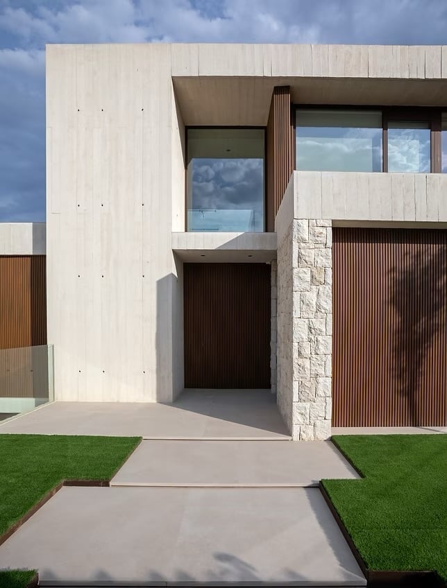 Modern white house exterior with geometric design, large windows, brown wooden accents, concrete walkway, and green lawn under blue sky