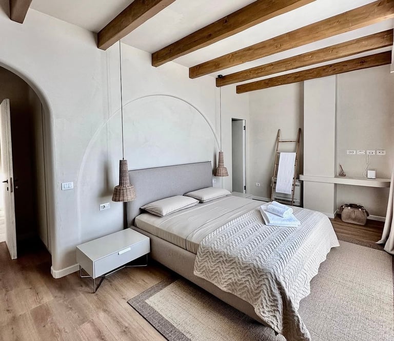 Modern bedroom with arched floor lamp, bed with gray upholstered headboard, wooden beams, white walls, and minimalist furniture