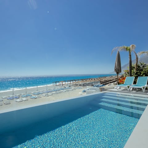 Beachfront pool with turquoise water, white lounge chairs, and palm trees overlooking Mediterranean Sea