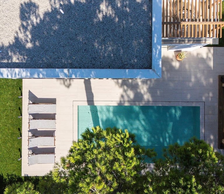 Aerial view of a backyard with a turquoise swimming pool, lounge chairs, lawn, and tree shadows on a blue wall