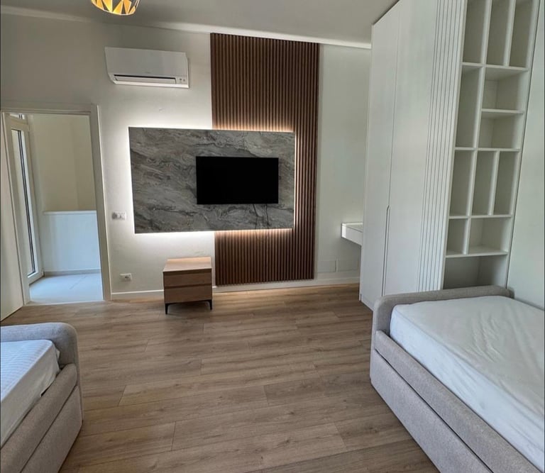 Modern bedroom with two beds, wall-mounted TV on textured accent wall, wooden flooring, and white shelving storage