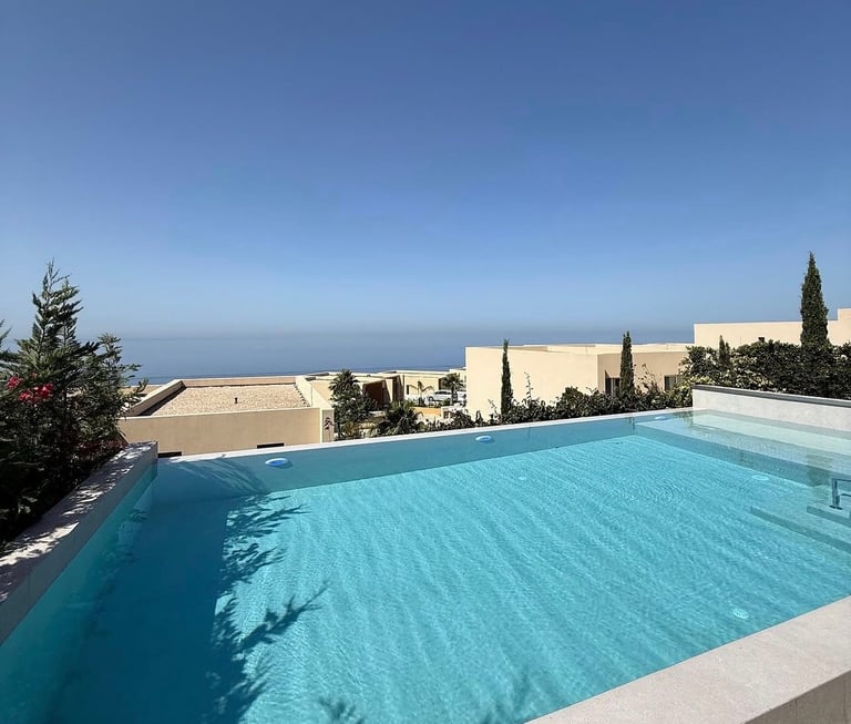 A bright turquoise swimming pool on a terrace with white stone buildings and cypress trees visible against a clear blue sky
