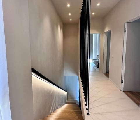 Modern hallway with recessed lighting, white walls, black vertical railing, and light tile flooring leading to multiple doors