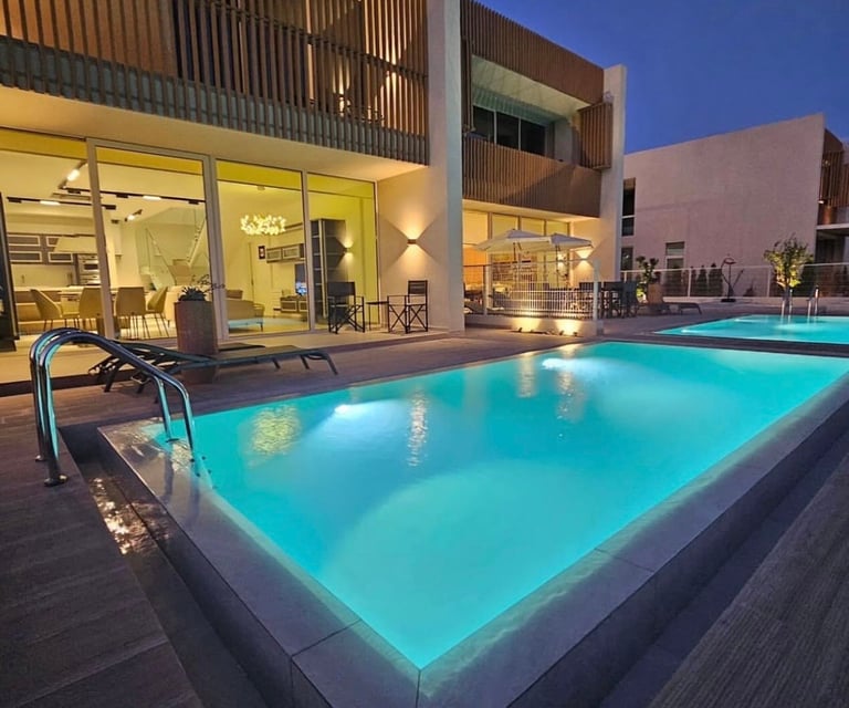 Illuminated swimming pool at dusk beside modern house with large windows, lounge chairs, and desert landscape at twilight