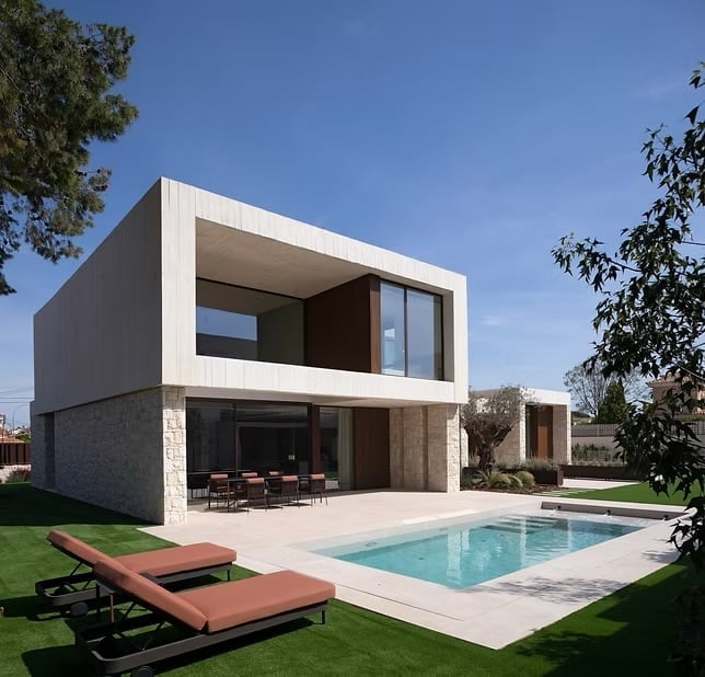 Modern two-story house with white and stone facade, pool, lounge chairs, and manicured lawn under clear blue sky