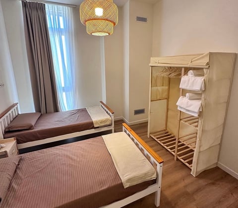 A modern hostel room with two single beds, a fabric wardrobe, woven pendant light, and floor-to-ceiling window with curtains