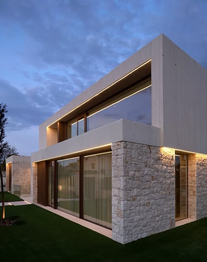 Modern cubic house with stone and white concrete exterior, illuminated at dusk, featuring large windows and geometric architecture.