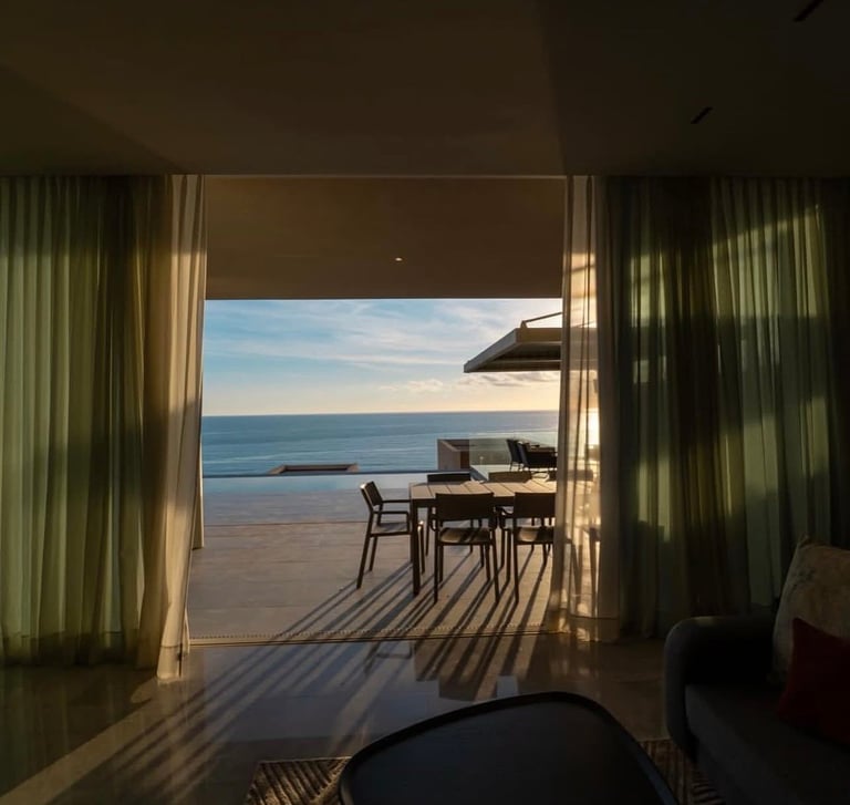 View from interior through open doors to seaside patio with dining furniture and ocean horizon at sunset