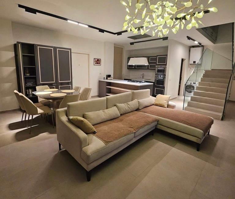 Modern living room showroom with cream sectional sofa, wooden accent, kitchen area, shelving, and staircase