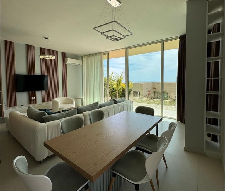 Modern open-plan living and dining room with gray sectional sofa, wooden dining table, floor-to-ceiling windows overlooking landscape, and built-in shelving