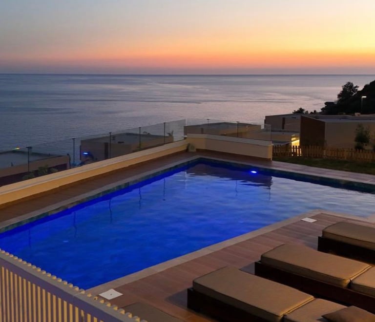 Illuminated swimming pool overlooking the ocean at sunset with loungers and coastal buildings