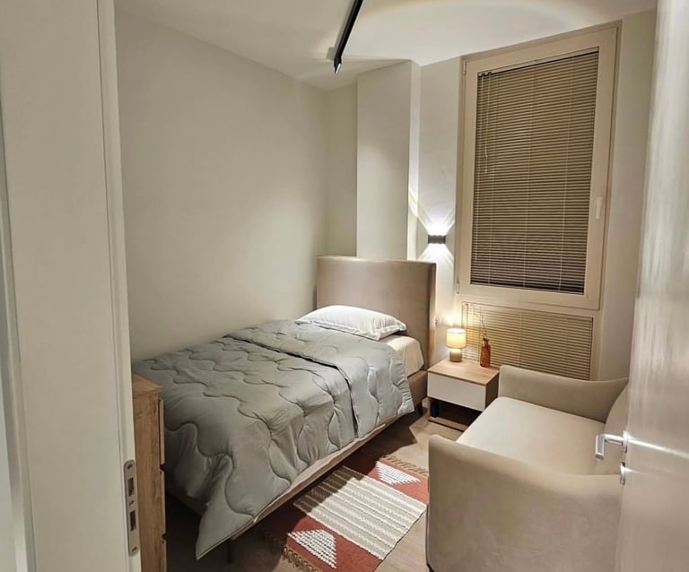 Minimalist bedroom with single bed, white bedding, wooden nightstand, and window with blinds overlooking urban view