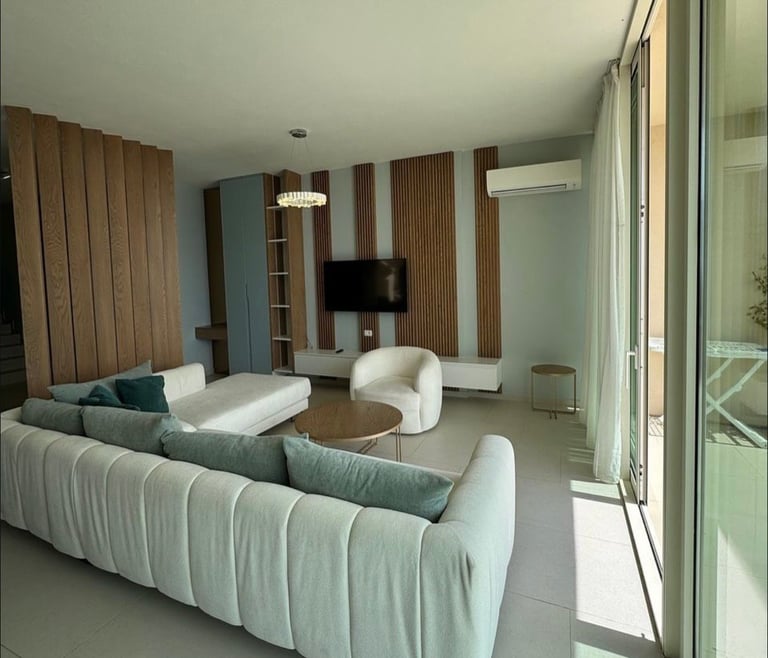 Modern living room with curved light blue sofa, white armchair, wooden TV wall unit, and glass doors leading to a balcony