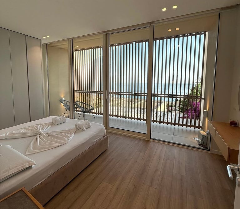 Modern bedroom with white bed, wooden floor, and floor-to-ceiling windows with vertical blinds overlooking a balcony.