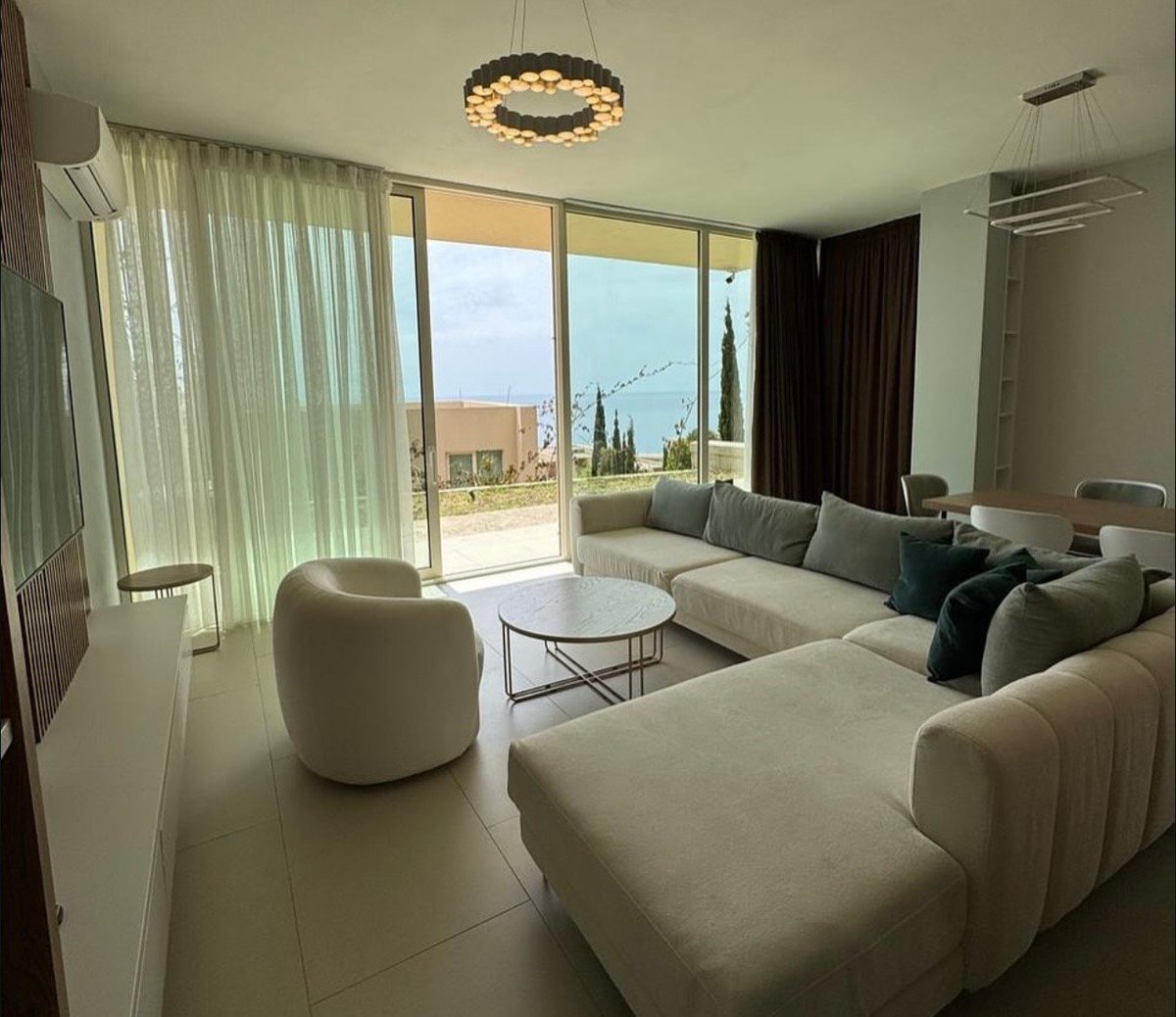 Modern living room with cream-colored sectional sofa, floor-to-ceiling windows, and contemporary lighting fixture overlooking a terrace.