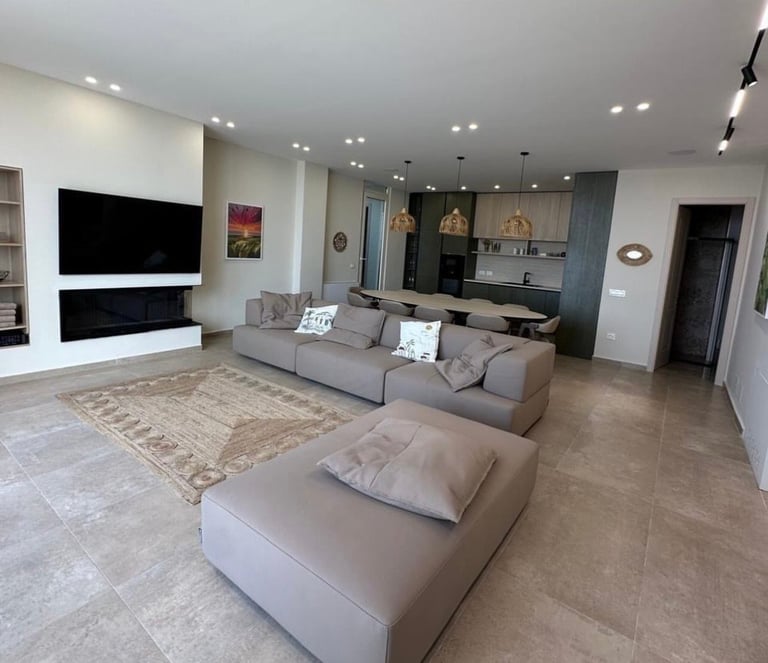 Modern open-concept living room with gray sectional sofa, wall-mounted TV, kitchen in background, and recessed ceiling lights