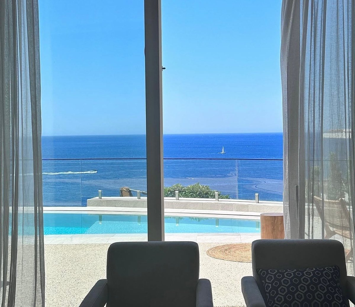 View from modern room with black chairs overlooking infinity pool and ocean horizon under clear blue sky
