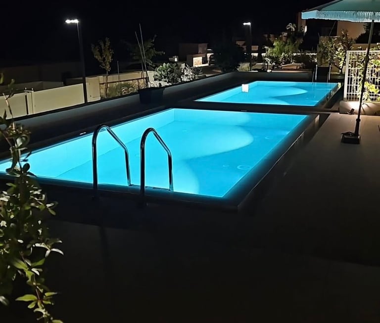 Two illuminated outdoor swimming pools at night with metal ladders, surrounded by landscaping and residential buildings