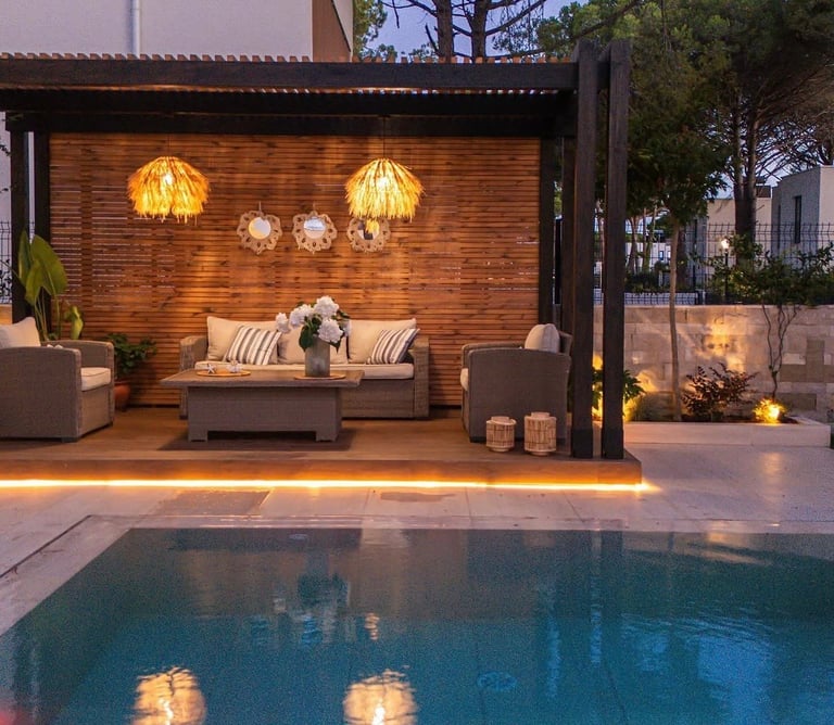 Modern poolside patio with illuminated lounge seating, pendant lights, and wood accent wall overlooking a blue swimming pool at dusk