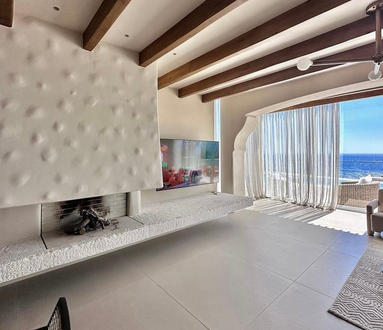 Modern luxury bedroom with ocean view, fireplace, exposed wood beams, and floor-to-ceiling windows overlooking the sea