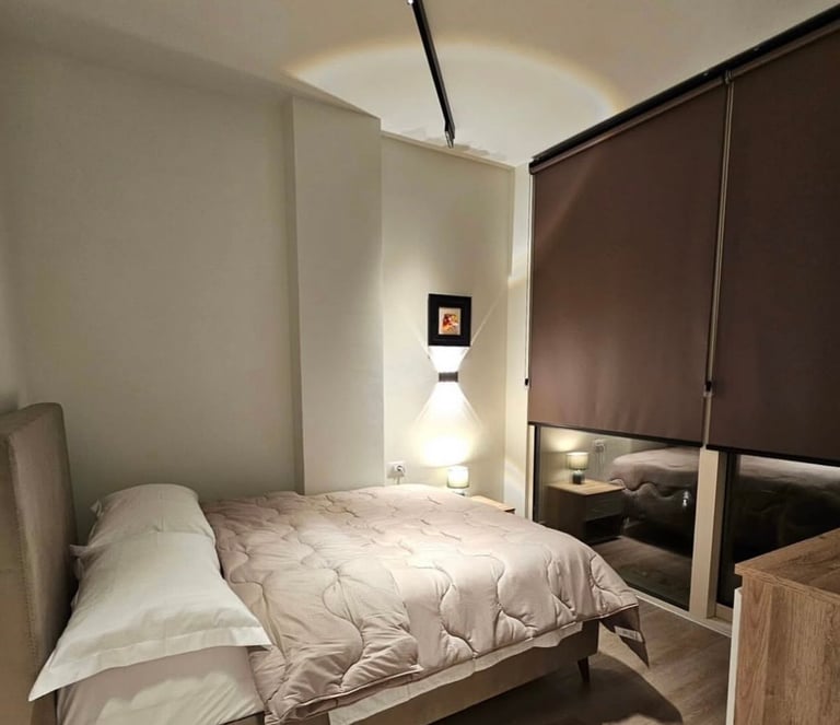 Minimalist bedroom with queen bed, white bedding, wall sconce lighting, brown roller shade, and wood nightstand