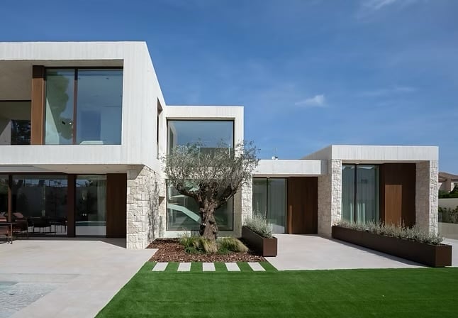 Modern white villa with geometric design, olive tree in courtyard, manicured lawn, and patio under clear blue sky