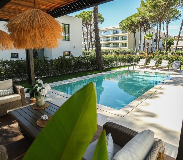 Luxury resort pool surrounded by lounge chairs, thatched umbrella, modern white buildings, and tall pine trees under blue sky