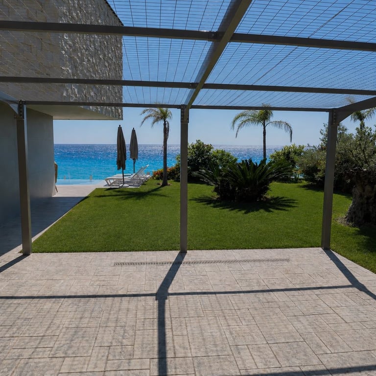 View through pergola to Mediterranean garden with green lawn, palm trees, and turquoise sea