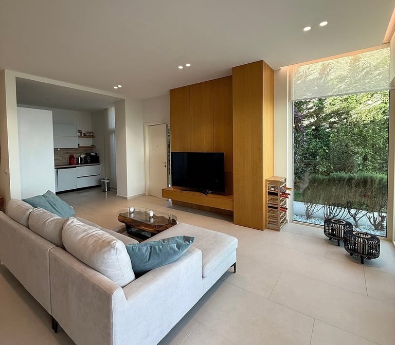Modern living room with beige sofa, wooden TV wall unit, open kitchen view, and large window overlooking garden