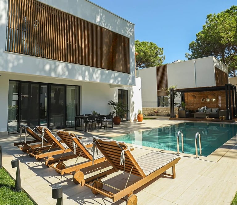 Modern white house with swimming pool, wooden sun loungers on deck, and slatted wooden privacy screens under clear blue sky
