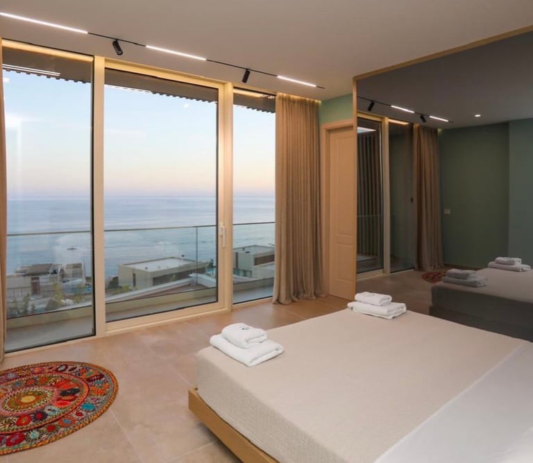 Modern bedroom with floor-to-ceiling windows overlooking a coastal seascape and sunset, wooden furniture, and minimalist decor