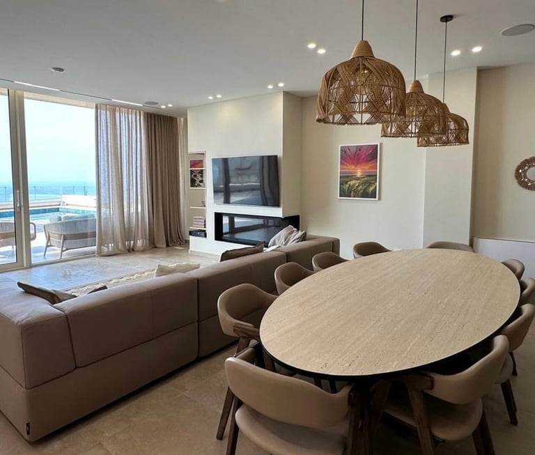 Modern open-concept living and dining area with oval table, beige seating, decorative pendant lights, and ocean view through floor-to-ceiling windows