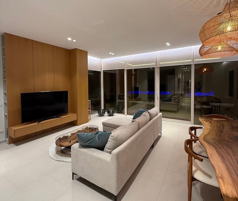 Modern living room with wooden TV wall, beige sofa, large glass windows overlooking city lights, and pendant lighting