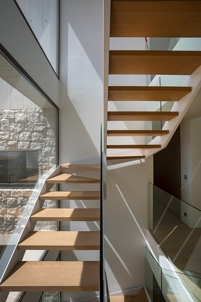 Modern spiral staircase with wooden treads, white walls, and a large window overlooking a stone exterior