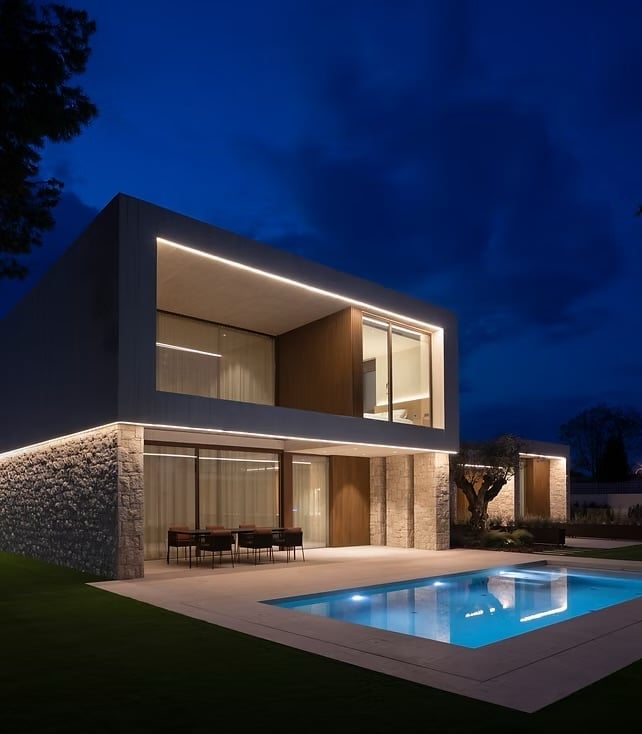 Modern two-story house with illuminated windows, stone lower level, and swimming pool at dusk